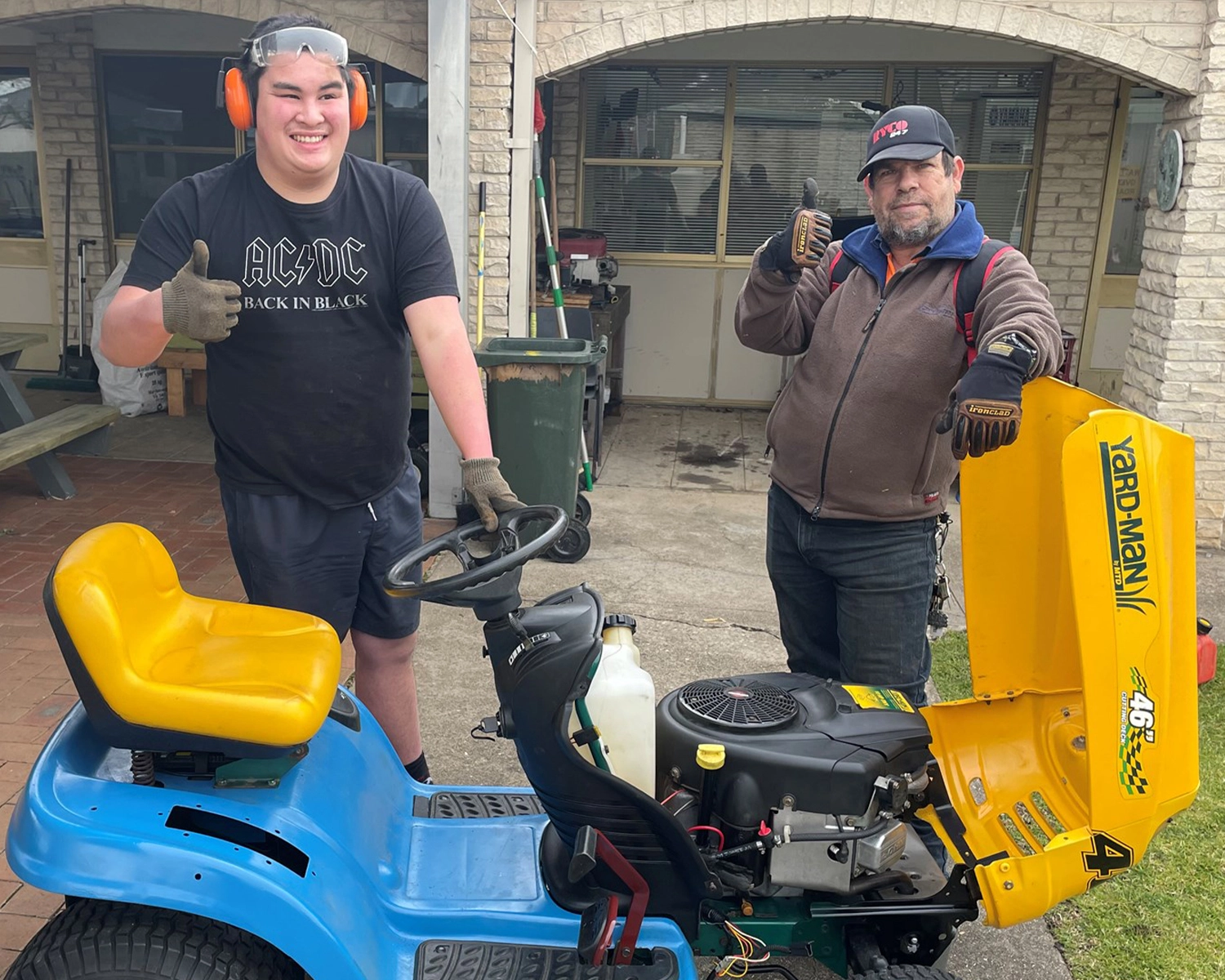 Two men wear safety clothing and stand next to a ride on lawnmower Disability supports Bairnsdale